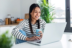 Smiling woman drinking coffee and researching cosmetic dentistry on laptop